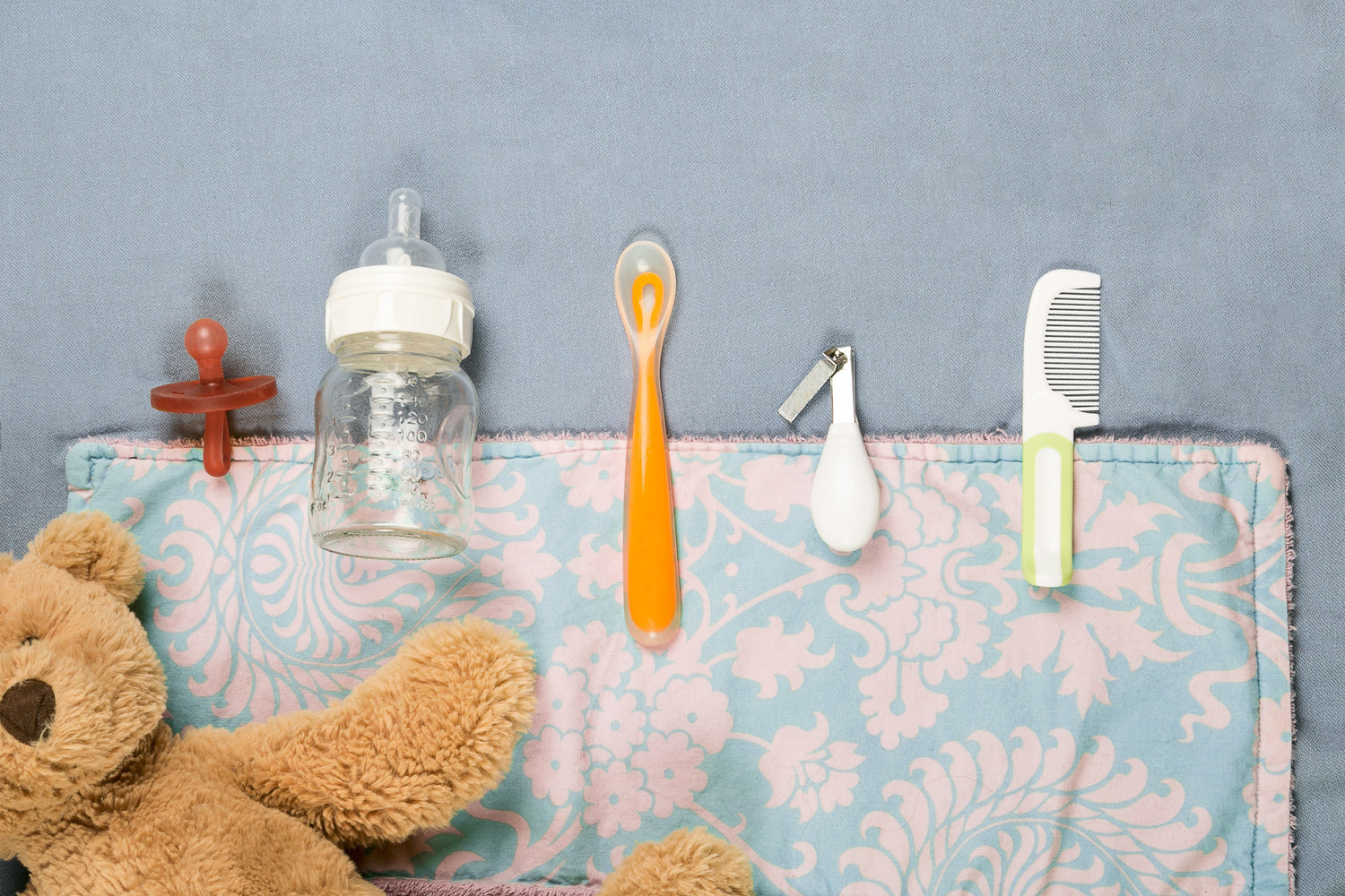 Flat lay of baby essentials including a baby bottle, pacifier, comb, nail clipper, spoon, and soft teddy bear on a decorative baby blanket.