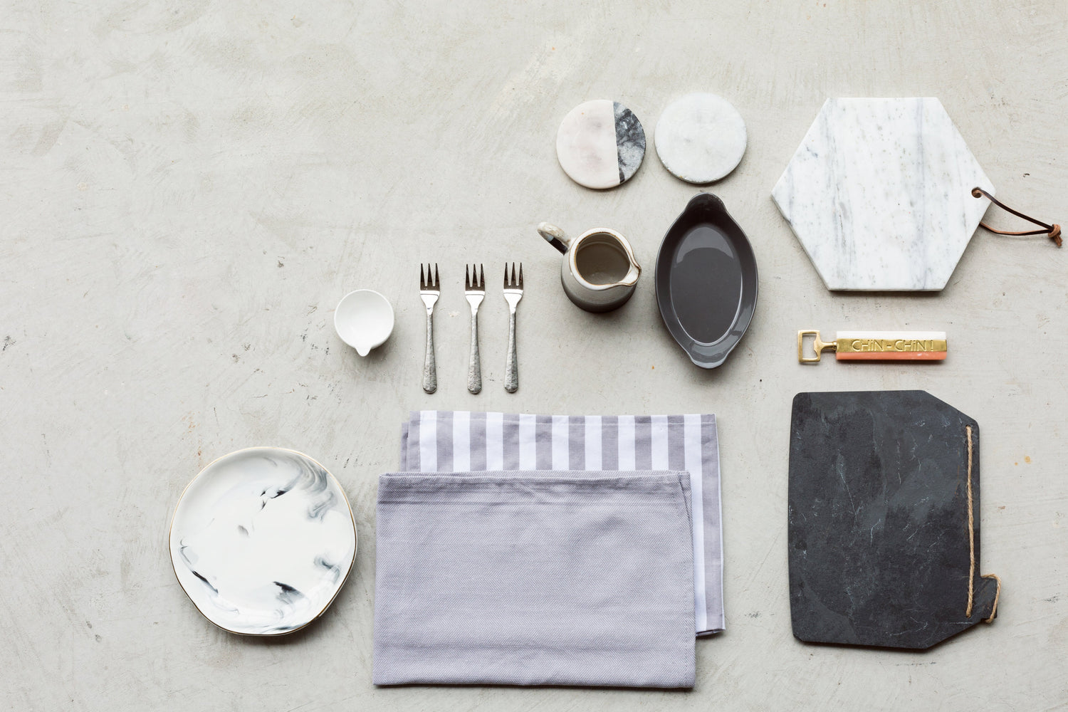 Elegant kitchen and dining accessories including marble coasters, flatware, napkins, ceramic plates, and a stylish bottle opener arranged on a light gray surface.