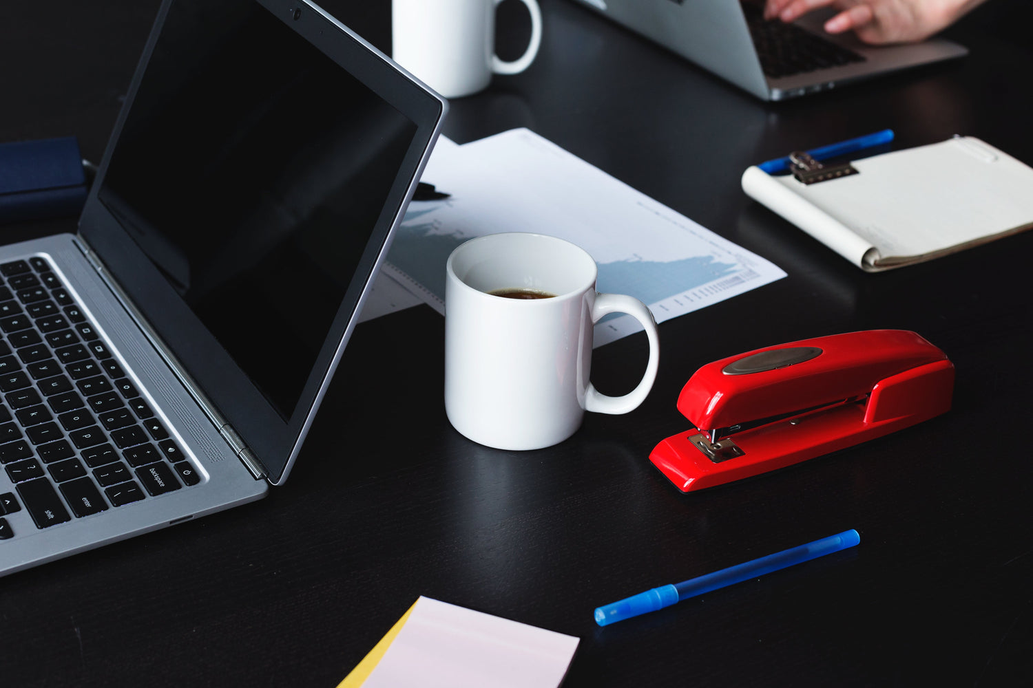 Office products on a desk including a laptop, red stapler, coffee mug, pen, and notepad, perfect for creating a productive workspace.