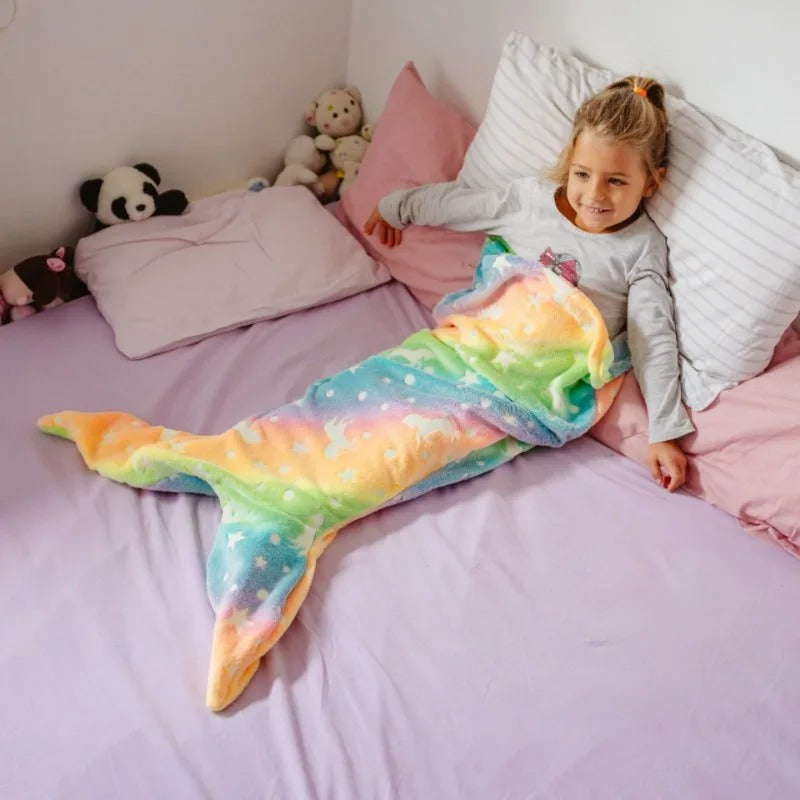 Child lying on bed wrapped in rainbow Mermaid Galaxy Glow Blanket smiling happily