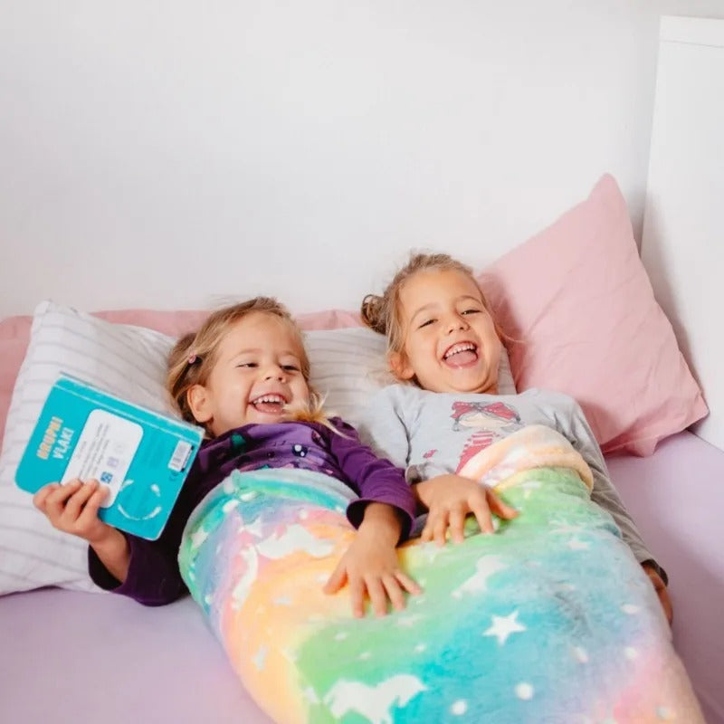 Two children laughing in bed while using rainbow Mermaid Galaxy Glow Blankets