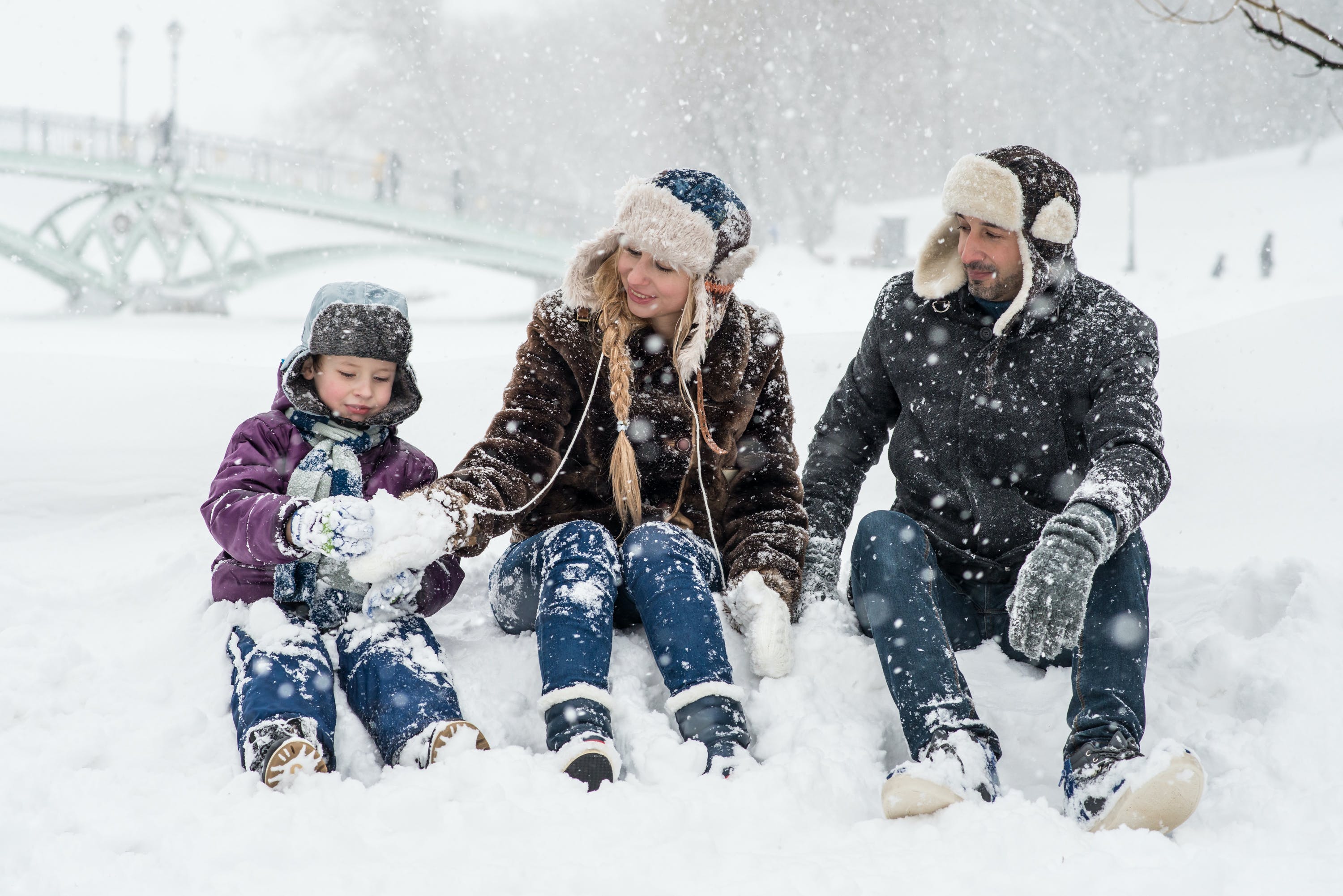 Happy family enjoying a snowy winter day outdoors, bundled up in warm coats and fur hats, making snowballs together in a park.